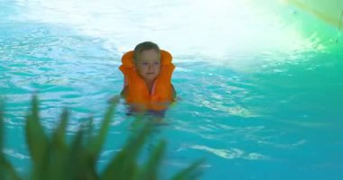 Turuncu can yeleği giyen genç bir çocuk havuzda yüzmekten hoşlanır. Parlak su ve yeşillik canlı, güvenli ve eğlenceli bir atmosfer yaratıyor..