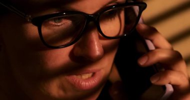 Gözlük takan bir kişi, sıcak aydınlatmayla aydınlatılmış bir telefon görüşmesiyle meşgul olur. Yakın çekim ifadeleri ve duyguları yakalar, samimi ve odaklanmış bir atmosfer yaratır..