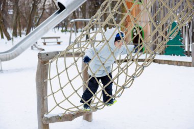 Kışın giyinmiş bir çocuk, karlı bir oyun bahçesinde ipe tırmanır. Sahne, arka planda bir slayt ve ağaçlarla kış boyunca açık hava oyun ve maceralarını yakalıyor..