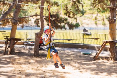 Kask takan ve koşum takımı takan genç bir çocuk ağaçlar ve doğa ile çevrili bir açık hava macera parkında heyecanlı bir ip yolculuğunun tadını çıkarıyor..
