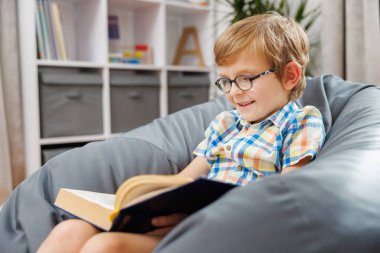 Gözlüklü genç bir çocuk rahat bir ev ortamında rahat bir fasulye koltukta otururken kitap okumaktan zevk alır. Arkaplanda bir kitaplık ve depolama kutuları yer alıyor.