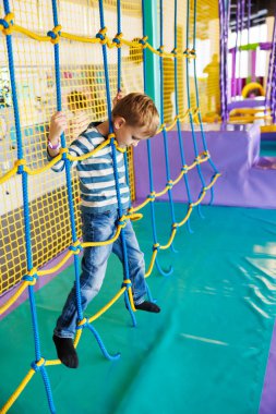 Kapalı bir oyun bahçesinde renkli bir halat ağında gezinen genç bir çocuk. Hareketli ortam fiziksel aktiviteyi ve oyunu, motor becerileri ve koordinasyonu geliştirmeyi teşvik eder..