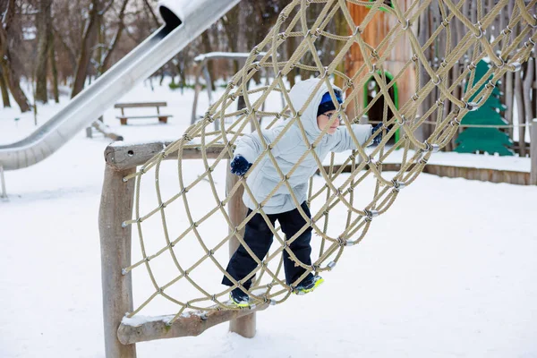 Kışın giyinmiş bir çocuk, karlı bir oyun bahçesinde ipe tırmanır. Sahne, arka planda bir slayt ve ağaçlarla kış boyunca açık hava oyun ve maceralarını yakalıyor..