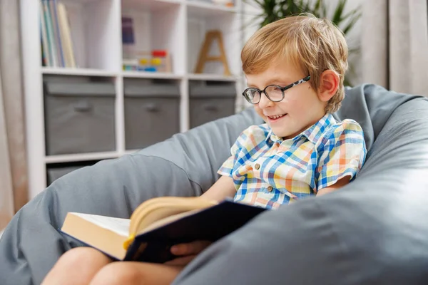 Gözlüklü genç bir çocuk rahat bir ev ortamında rahat bir fasulye koltukta otururken kitap okumaktan zevk alır. Arkaplanda bir kitaplık ve depolama kutuları yer alıyor.