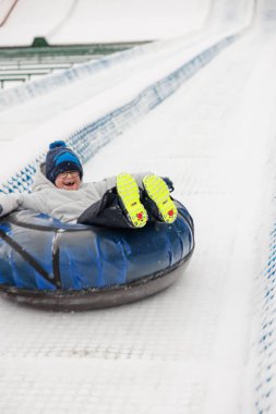 Kışın giyinmiş bir çocuk karlı bir yamaçta neşeyle kar tüneline biner. Tüpün parlak renkleri ve çocuk ayakkabıları beyaz karla tezat oluşturuyor, eğlenceli bir kış aktivitesi yakalıyorlar..
