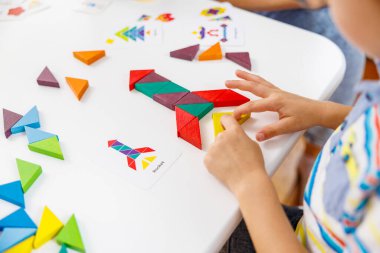 Bir çocuk beyaz bir masada renkli bir geometrik bulmacayla meşgul olur, desen kartıyla eşleştirmek için tahta şekiller kullanır. Bu etkinlik yaratıcılığı ve bilişsel gelişimi teşvik eder..