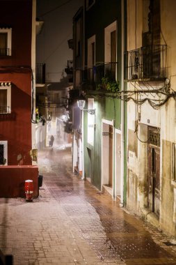 A vibrant, narrow street scene with brightly colored red and green buildings on either side, where the wet cobblestone reflects the dim light, leading the eye into the foggy, atmospheric distance where a lone figure walks.