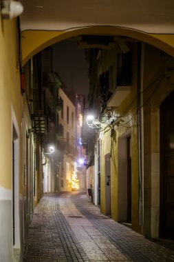 A warm, inviting photograph taken through a yellow-painted archway leading into a narrow, wet alley in the old town of Villajoyosa. The light from a distant, blurred Christmas tree and streetlights pierces the misty air, highlighting the cobblestone 