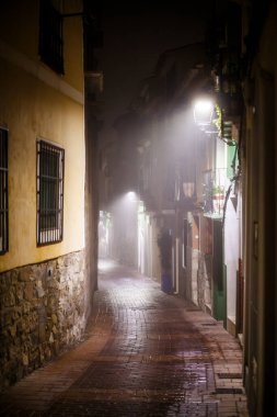 A deeply atmospheric photograph capturing a narrow, cobbled alleyway in Villajoyosa on a foggy night. The warm, yellow light from a street lamp illuminates the wet, brick-paved ground, reflecting the light and enhancing the contrast with the shadows 