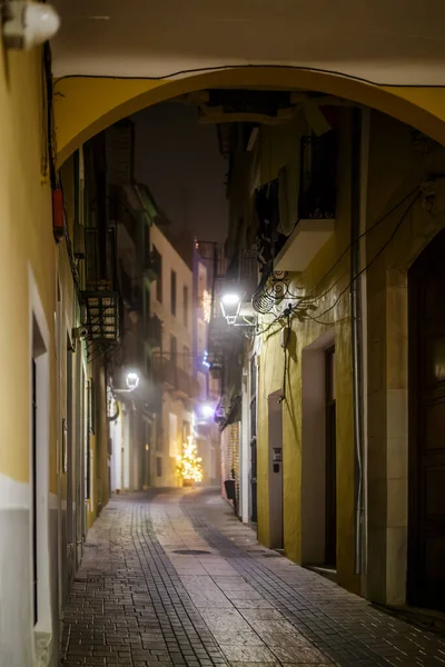 A warm, inviting photograph taken through a yellow-painted archway leading into a narrow, wet alley in the old town of Villajoyosa. The light from a distant, blurred Christmas tree and streetlights pierces the misty air, highlighting the cobblestone 