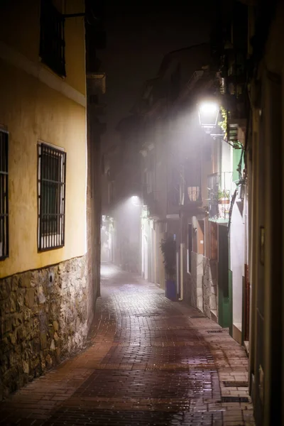 A deeply atmospheric photograph capturing a narrow, cobbled alleyway in Villajoyosa on a foggy night. The warm, yellow light from a street lamp illuminates the wet, brick-paved ground, reflecting the light and enhancing the contrast with the shadows 