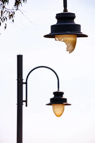 İki sokak lambasının ön plandaki kırık cam küreyi aydınlatan parlak beyaz-sarı gökyüzüne karşı dikey bir çekim..