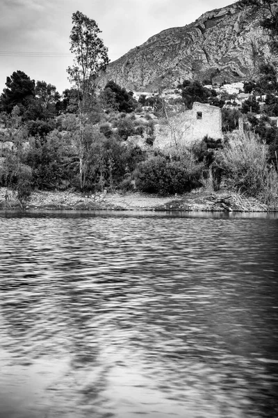 Amadorio Reservoir 'ın kıyılarının siyah ve beyaz bir görüntüsü, çalılık bir yamaç ve desenli bir dağ zeminine karşı önemli bir harabe bina seti içeriyor. Ön plandaki su dinamiktir, açık bir dalga etkisi gösterir..