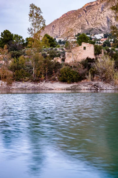Alacakaranlıkta ya da şafakta Amadorio Barajı 'nın renkli bir fotoğrafı. Sıcak bir ışık kıyıdaki harap olmuş bir yapıyı aydınlatıyor. Ön plandaki su dalgalanmalar ve mavi ve yeşil tonların karışımıyla canlıdır..