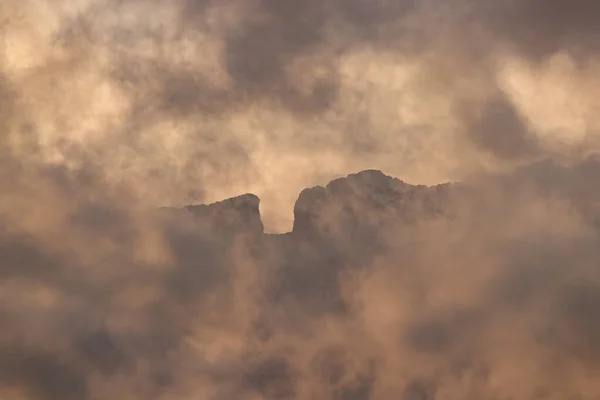 Benidorm 'un üzerinde gün batımında sıcak, fırtınalı bulutların arasından zar zor görünen engebeli bir dağın dramatik, yakın çekim görüntüsü..