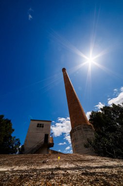 Yüksek tuğlalı bacanın güçlü, alçak açılı görüntüsü ve güneşten sırılsıklam olmuş parlak mavi bir gökyüzü tarafından çerçevelenmiş harap olmuş beton bir bina. Küçük, ıssız sarı bir çiçek çatlamış ön plandaki Symb 'dan esnek bir şekilde büyür.