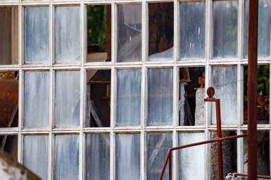 Yatay çekim, toz ve kirle kaplı birçok küçük pencere penceresinin detaylı ızgarasına odaklanmış. Birkaç pencere eksik veya kırık. İçindeki karanlık ve paslı makineleri ortaya çıkarıyor..