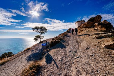 İki yürüyüşçü, parlak, mavi Akdeniz 'in bir zeminine karşı tarihi Malladeta kulesine (veya benzer bir gözcü yapısı) doğru tozlu bir kıyı yoluna yükselir ve parlak, güneşli bulutlu bir gökyüzü.