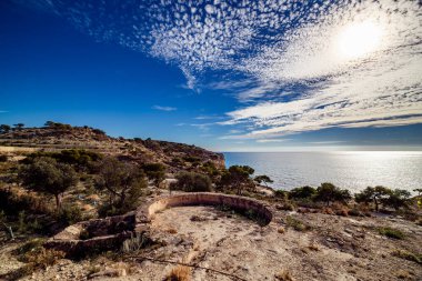 La Malladeta 'dan bir manzara dairesel bir taşın kalıntılarını gösteriyor, muhtemelen kadim bir gözetleme noktası, parıldayan Akdeniz ve kıyı şeridi mavi, bulutlu bir gökyüzü altında uzanıyor..