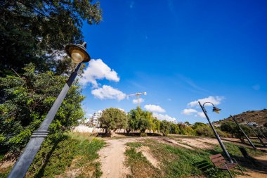 Toprak yolu ve iki sokak lambası olan bir park alanından geniş açılı bir manzara, yeşil bir manzaraya bakan bir bina vincinin canlı mavi gökyüzüne karşı yükseldiği yer..