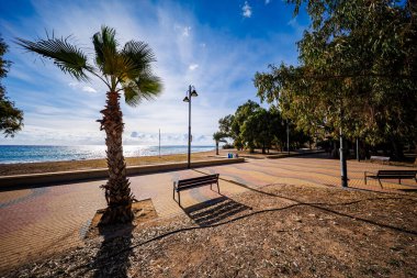 Renkli gezinti güvertesi boyunca, ön planda büyük bir palmiye ağacı ve ikiz başlı bir sokak lambasının yanında tek kişilik bir bank bulunan biraz merkezden uzak bir manzara. Sahne, gözleri parıldayan denize ve güneşli plaja yönlendirir..