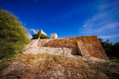 Bir yamaçtaki eski bir taş istinat duvarının alçak açılı çekimi, muhtemelen Roma 'nın Allon yerleşim yerinin yakınında. Küçük, beyaz bir yapı, duvarın üzerinde, açık mavi bir gökyüzüne karşı duruyor..