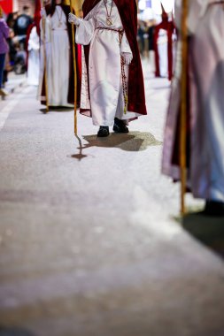 Beyaz bir tunik ve kırmızı kadife pelerinli bir tövbekârın alt yarısına odaklanan yakın plan bir çekim, kaldırımda uzun, sembolik bir gölge oluşturan ahşap bir asa tutuyor..