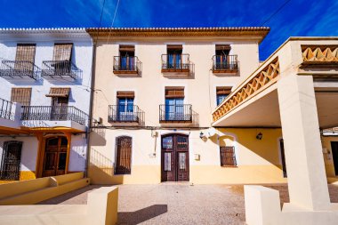 Üç katlı klasik bir Akdeniz binasının geniş açılı bir görüntüsü dövülmüş demir balkonları ve ahşap panjurları olan, güneşli bir kasaba meydanında yer alan.