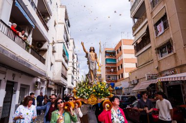 Villajoyosa, Alicante, İspanya. 2025-04-20. Yükselen İsa 'nın yol ayrımında yüzdüğünü gösteren yüksek açılı bir sokak manzarası. Ön planda bir kadın, sepetten yaprakları ya da kağıtları heykele doğru fırlatıyor..