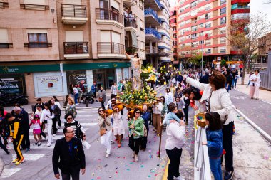 Villajoyosa, Alicante, İspanya. 2025-04-20. Yükselen İsa 'nın yol ayrımında yüzdüğünü gösteren yüksek açılı bir sokak manzarası. Ön planda bir kadın, sepetten yaprakları ya da kağıtları heykele doğru fırlatıyor..