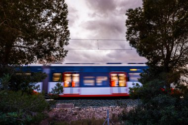 Geçen bir trenin iç ışıkları, karanlık, bulutlu bir gökyüzüne ve Akdeniz bitkisinin siluetlerine karşı canlı renk çizgileri oluşturur..