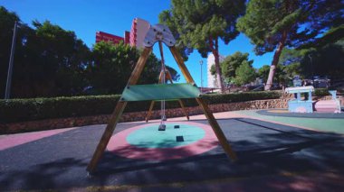 Geniş açılı, halka açık bir oyun parkında, arka planda yumuşak güvenlik zemini ve yüksek apartman binaları ve açık mavi gökyüzünün altında çam ağaçları bulunan modern bir ahşap salıncak görüntüsü..