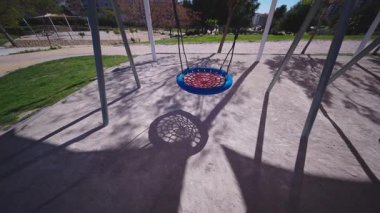 Benidorm 'da aydınlık bir günde büyük bir gölgeyle gölgelenmiş, boş bir oyun bahçesinde asılı mavi ve kırmızı yuvarlak bir yuva salıncağı geniş bir çekim.