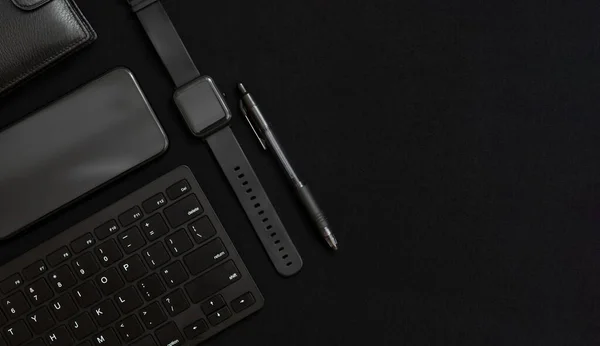 Flat lay composition with black pen, computer keyboard, smart watch, smartphone and leather wallet on dark black surface. Business concept. Business people items. Free space for text. Low key photo. Stationery. Items of successful people.