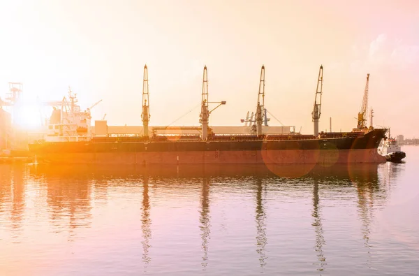 Bulk carrier ship in the port on loading. Bulk cargo ship under port crane bridge. Ship in seaport at sunset. Sun rays and glare. Toned photo.