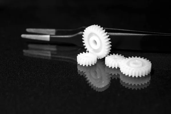 Several white plastic gears and tweezers on a black mirror background. Mechanical repair. Fix. Reflection. Repair shop. Cogwheels. Elements of a large mechanism. Harmonious work. Business success. Blurred background.