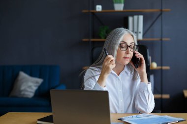 Gülümseyen kıdemli gri saçlı güzel iş kadını ofiste telefonda konuşuyor. İş yerinde oturan, gözlüklü, çekici, olgun bir kadının iş portresi. Konuşma, müzakere