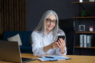 Kıdemli gri saçlı güzel kadın akıllı telefon kullanıyor ve gülümsüyor. Mutlu iş kadını cep telefonu uygulamaları kullanıyor, mesaj atıyor, internette geziniyor, internetten alışveriş yapıyor. Mobil aygıtları olan olgun insanlar