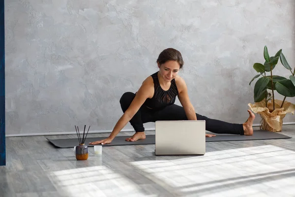 Young sporty woman watching online yoga lessons on laptop. Beautiful girl practicing side lunge pose, skandasana. Yoga postures, meditation, relaxation, stress management, wellbeing and health
