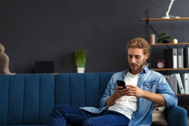 Akıllı telefon kullanıp gülümseyen kıvrımlı adam. Mutlu adam cep telefonu uygulamaları kullanıyor, mesaj yazıyor, internette geziniyor, akıllı telefona bakıyor, evde oturuyor. Mobil aygıtlarla çalışan gençler