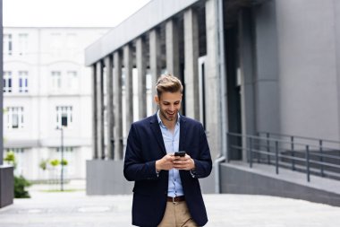 Akıllı telefon kullanan ve gülümseyen yakışıklı bir iş adamı. Mutlu genç adam cep telefonu uygulamaları kullanıyor, mesaj atıyor, internette geziniyor, akıllı telefona bakıyor. Mobil aygıtlarla çalışan gençler