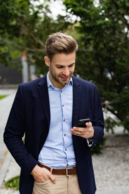 Akıllı telefonu tutan ve gülümseyen yakışıklı bir ofis çalışanı. Mutlu genç adam cep telefonu uygulamaları kullanıyor, mesaj atıyor, internette geziniyor, akıllı telefona bakıyor. İş merkezinin yakınında kahve molası.