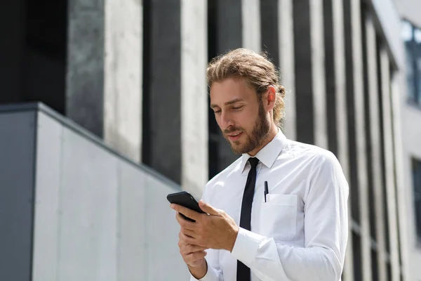 Портрет успешного молодого бизнесмена. Кудрявый мужчина в белой рубашке с телефоном на фоне современного бизнес-центра — стоковое фото