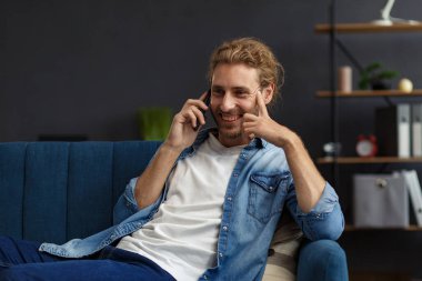 Akıllı telefon kullanıp gülümseyen kıvrımlı adam. Mutlu adam cep telefonu uygulamaları kullanıyor, mesaj yazıyor, internette geziniyor, akıllı telefona bakıyor, evde oturuyor. Mobil aygıtlarla çalışan gençler