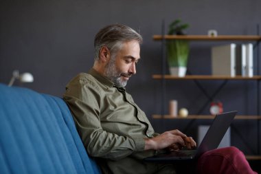 Merkez ofiste dizüstü bilgisayarla çalışan kıdemli bir adam. Kanepede oturan yakışıklı gri saçlı müdürün portresi. Çevrimiçi, çevrimiçi kurslar. Çevrimiçi eğitim, uzaktan çalışma