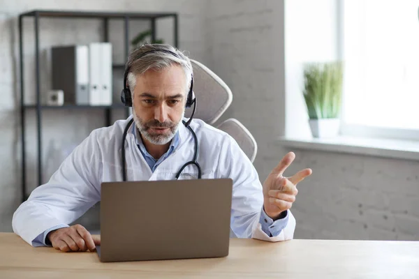 Portrait d'un médecin chevelu aux cheveux gris dans son bureau utilisant un ordinateur portable pour le chat vidéo avec un patient. Consultation en ligne avec le médecin pour les diagnostics et les recommandations de traitement. Concept de télésanté — Photo