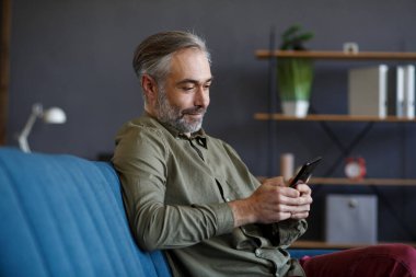 Gülümseyen gri saçlı adam cep telefonu uygulamaları kullanıyor, mesaj atıyor, internette geziniyor, internetten alışveriş yapıyor. Akıllı telefona bakmak, gülümsemek, evde oturmak. Mobil aygıtları olan olgun insanlar