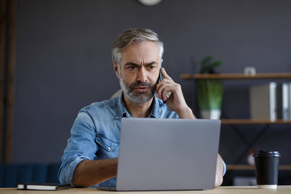 Senior grey-haired businessman talking on the phone in office. Conversation with business partners. Business portrait of handsome mature man sitting at workplace. Communication, negotiation