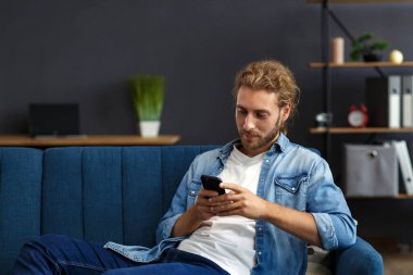 Akıllı telefon kullanıp gülümseyen kıvrımlı adam. Mutlu adam cep telefonu uygulamaları kullanıyor, mesaj yazıyor, internette geziniyor, akıllı telefona bakıyor, evde oturuyor. Mobil aygıtlarla çalışan gençler