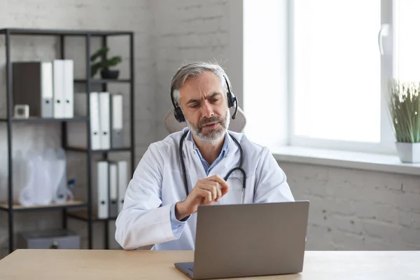 Portrait d'un médecin chevelu aux cheveux gris dans son bureau utilisant un ordinateur portable pour le chat vidéo avec un patient. Consultation en ligne avec le médecin pour les diagnostics et les recommandations de traitement. Concept de télésanté — Photo